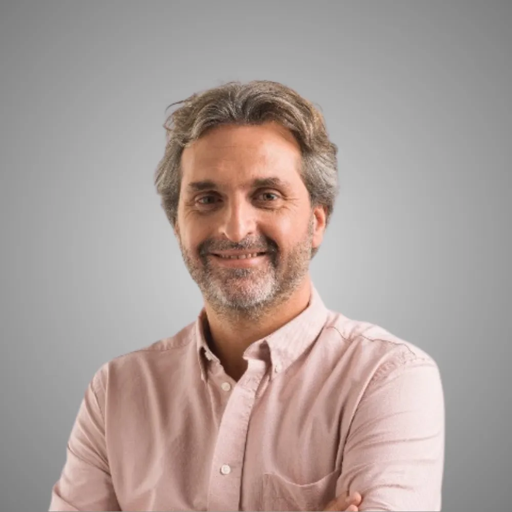Homme souriant aux cheveux gris portant une chemise rose sur fond gris uni.