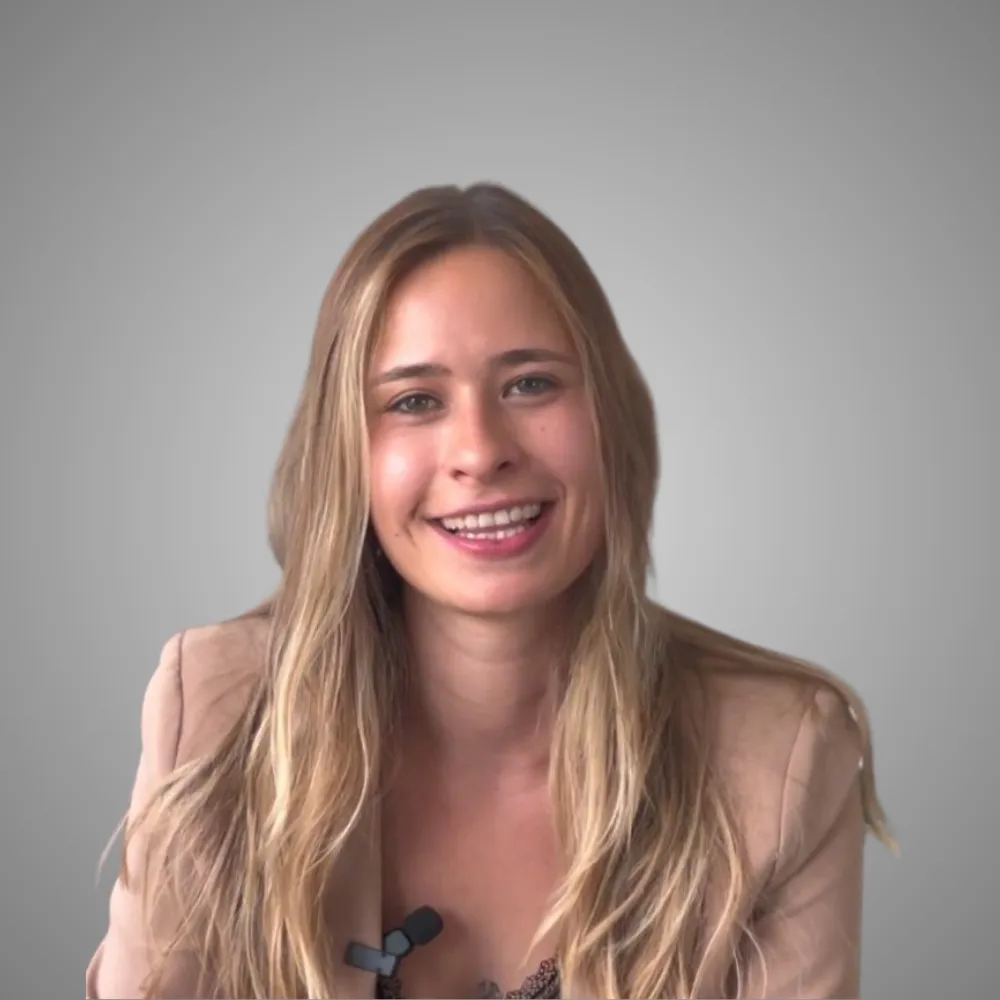 Jeune femme souriante aux cheveux blonds longs portant une veste beige sur fond gris.