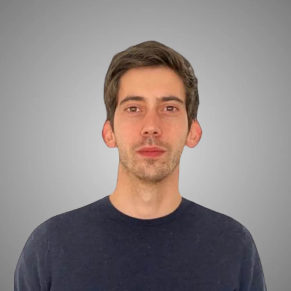 Homme avec cheveux bruns courts et pull bleu foncé devant un fond gris uni.
