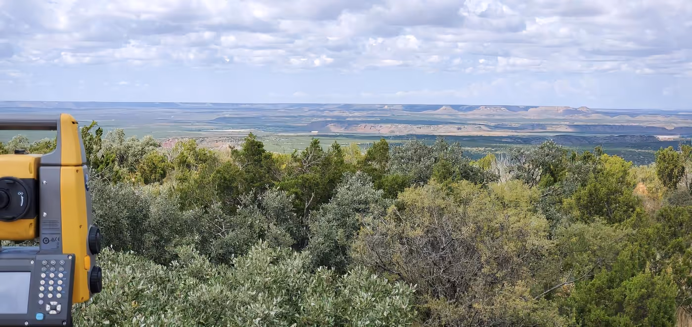 Surveying equipment positioned overlooking dense green shrubbery with expansive flat-topped mesas in the distant horizon under a cloudy sky.