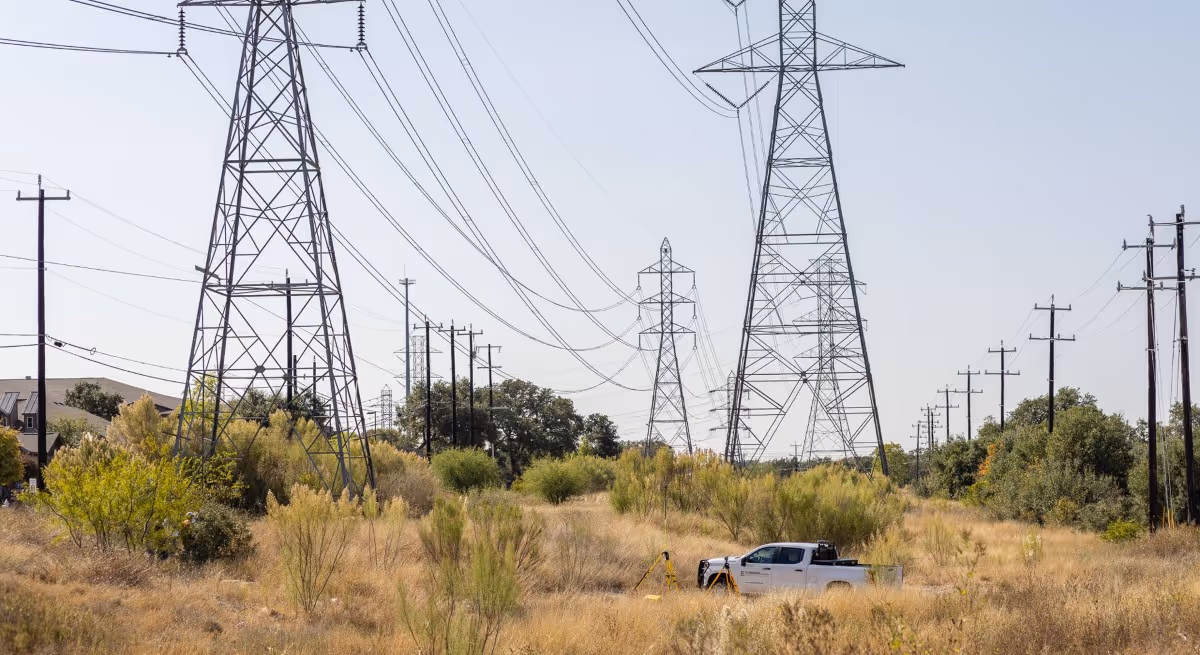 White pickup truck and surveying equipment in a dry grassy field beneath large electrical transmission towers and power lines.