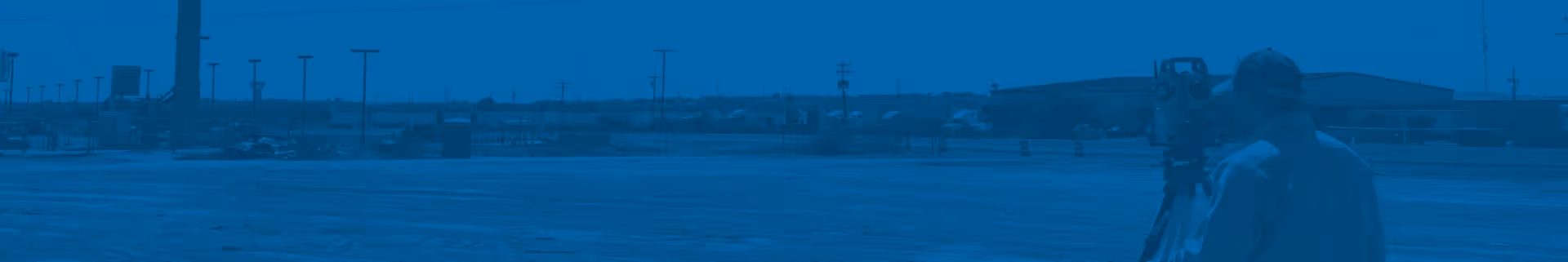 Construction worker using surveying equipment in an open cleared land area near buildings and power lines.
