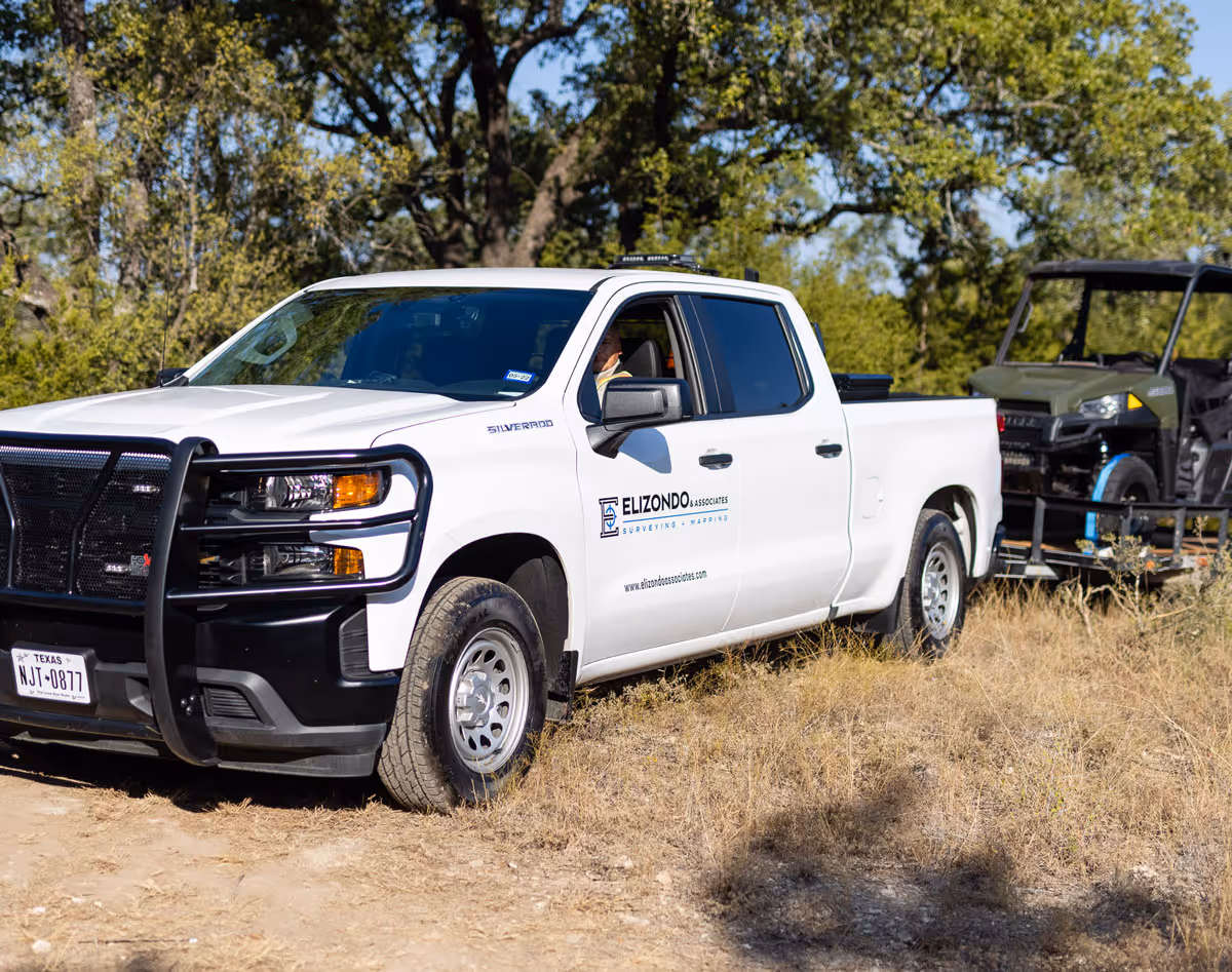 White truck with Elizondo & Associates logo parked on dry grass, towing a trailer carrying an off-road vehicle.