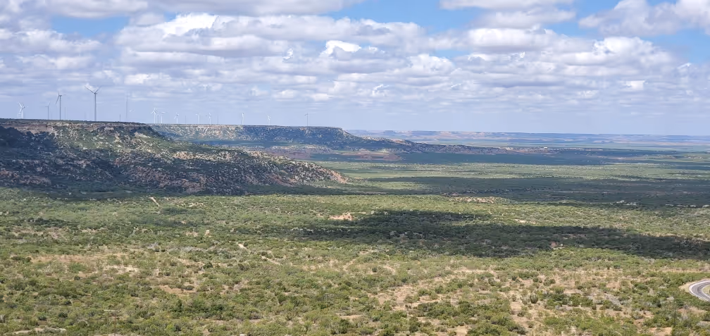 Wide landscape view of green scrubland with a ridge in the distance and wind turbines along the ridge under a partly cloudy sky.