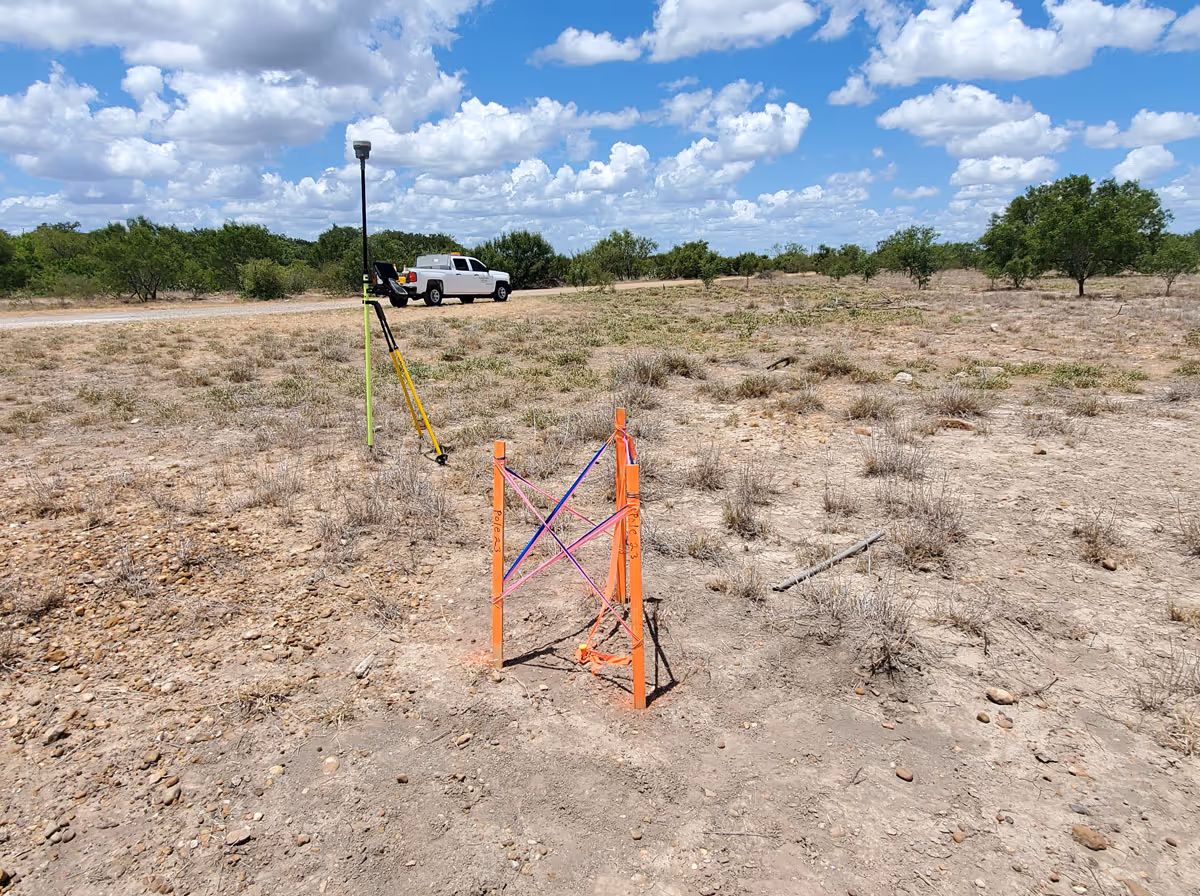 Surveying equipment set up in a dry, rocky field with a white pickup truck and trees in the background under a partly cloudy sky.
