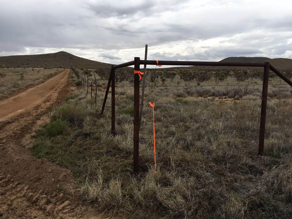 Rusty metal fence and barbed wire bordering a dirt road in a dry, grassy landscape under a cloudy sky.