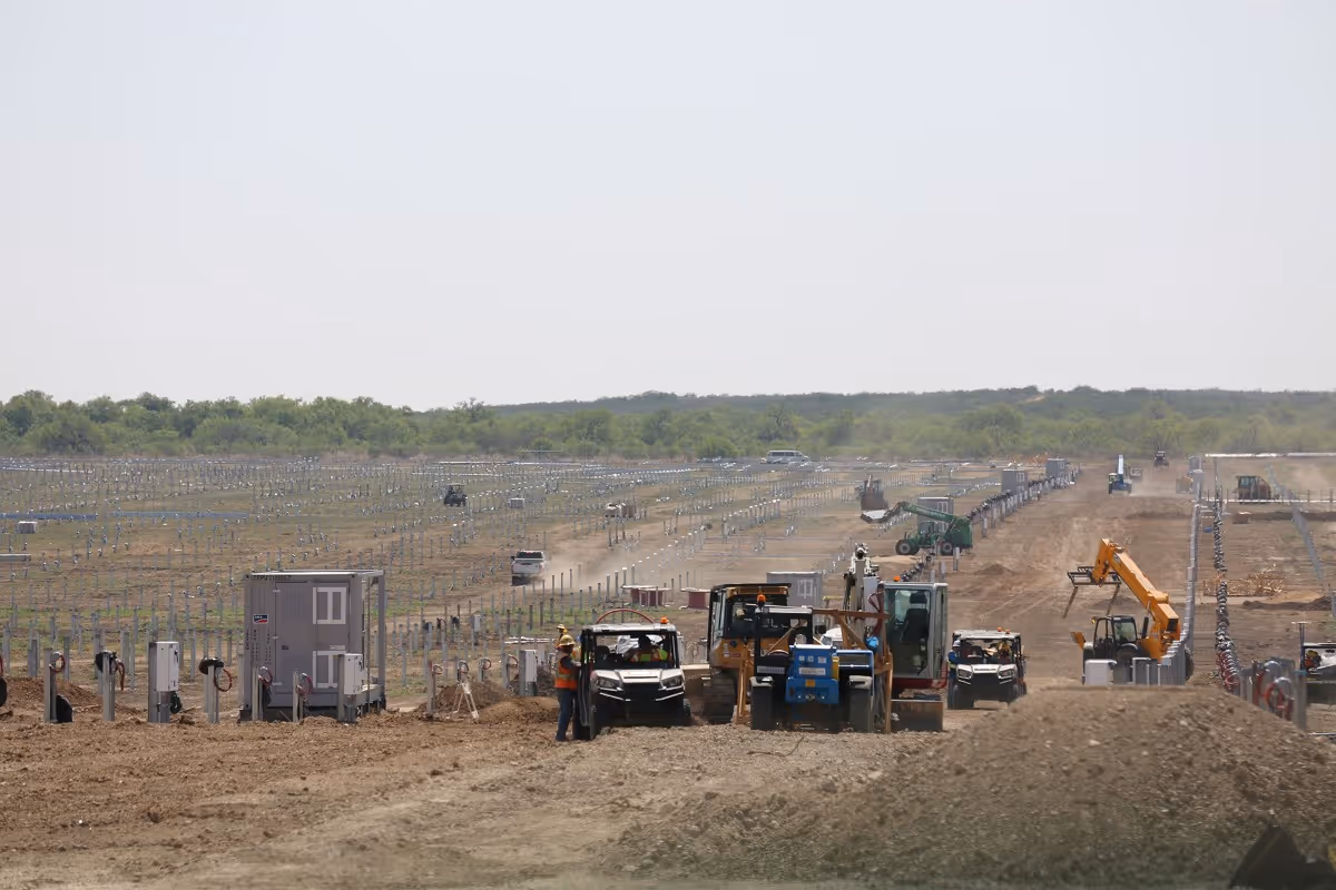Construction site with heavy machinery and workers installing solar panel mounts in a large field.