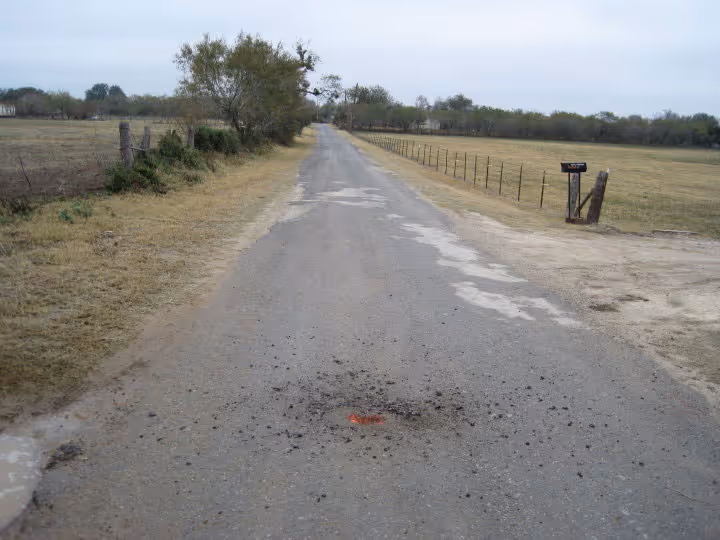 Rural paved road with an orange survey marker surrounded by disturbed gravel in the center, flanked by fields and fences under a cloudy sky.