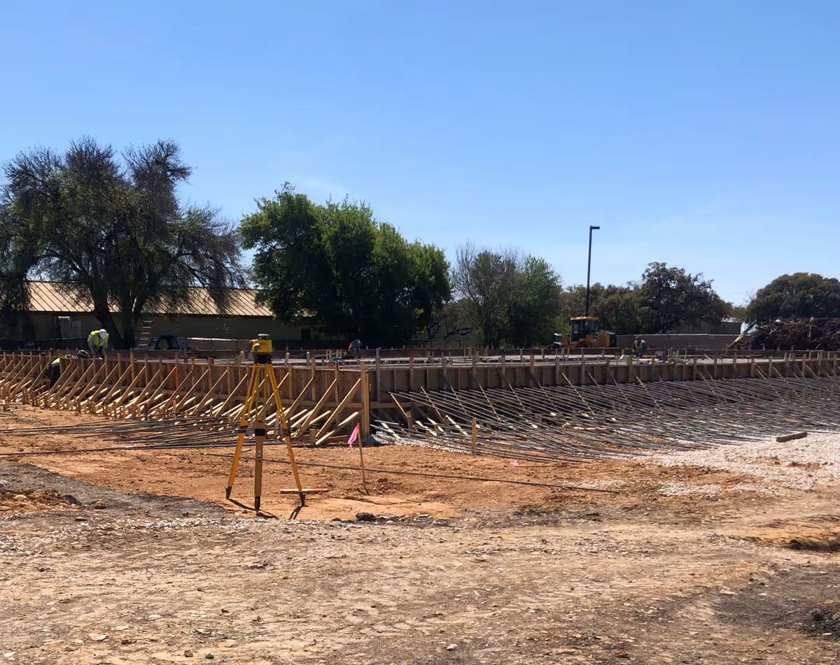 Construction site with wooden formwork, rebar reinforcement, and a surveying tripod under clear blue sky.