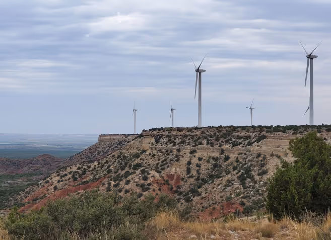 Wind turbines on a ridgeline in a semi-arid landscape with shrubs and a cloudy sky.