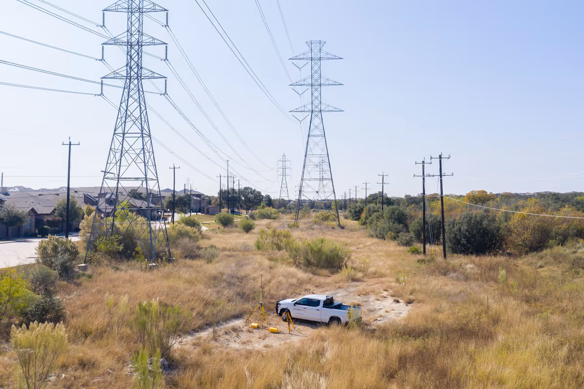 White pickup truck with surveying equipment set up in a clearing beneath power lines and transmission towers.