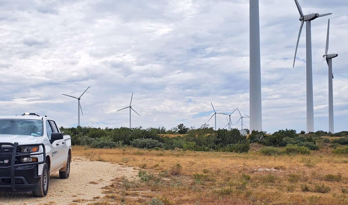 White pickup truck parked on dirt path near several large wind turbines in a grassy field under a cloudy sky.