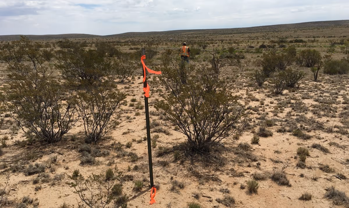 Person in orange safety vest standing in a dry, shrub-covered desert landscape with a stake wrapped with orange ribbon in the foreground.