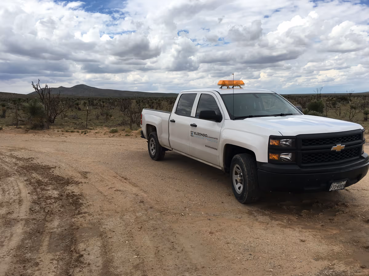 White Chevrolet pickup truck with orange light on top parked on a dirt road in a desert landscape under a cloudy sky.