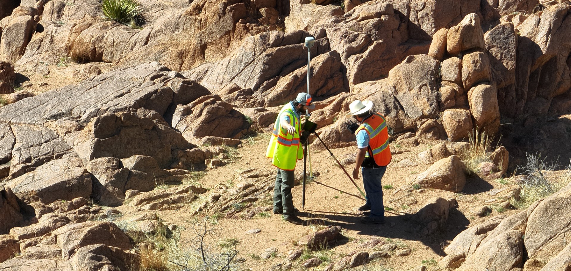 Two surveyors in safety vests measuring rocky terrain with surveying equipment.