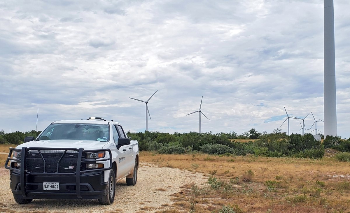 Survey vehicle parked on dirt road near wind turbines in rural landscape.