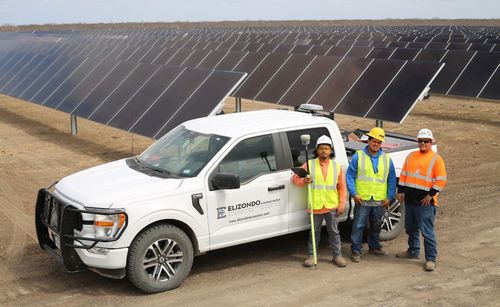 Three workers in safety gear standing beside a white survey truck near solar panel arrays.