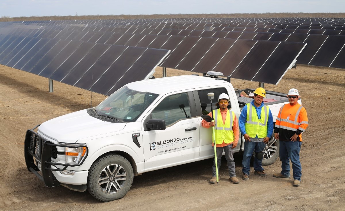 Three workers in safety gear standing beside a white survey truck near solar panel arrays.