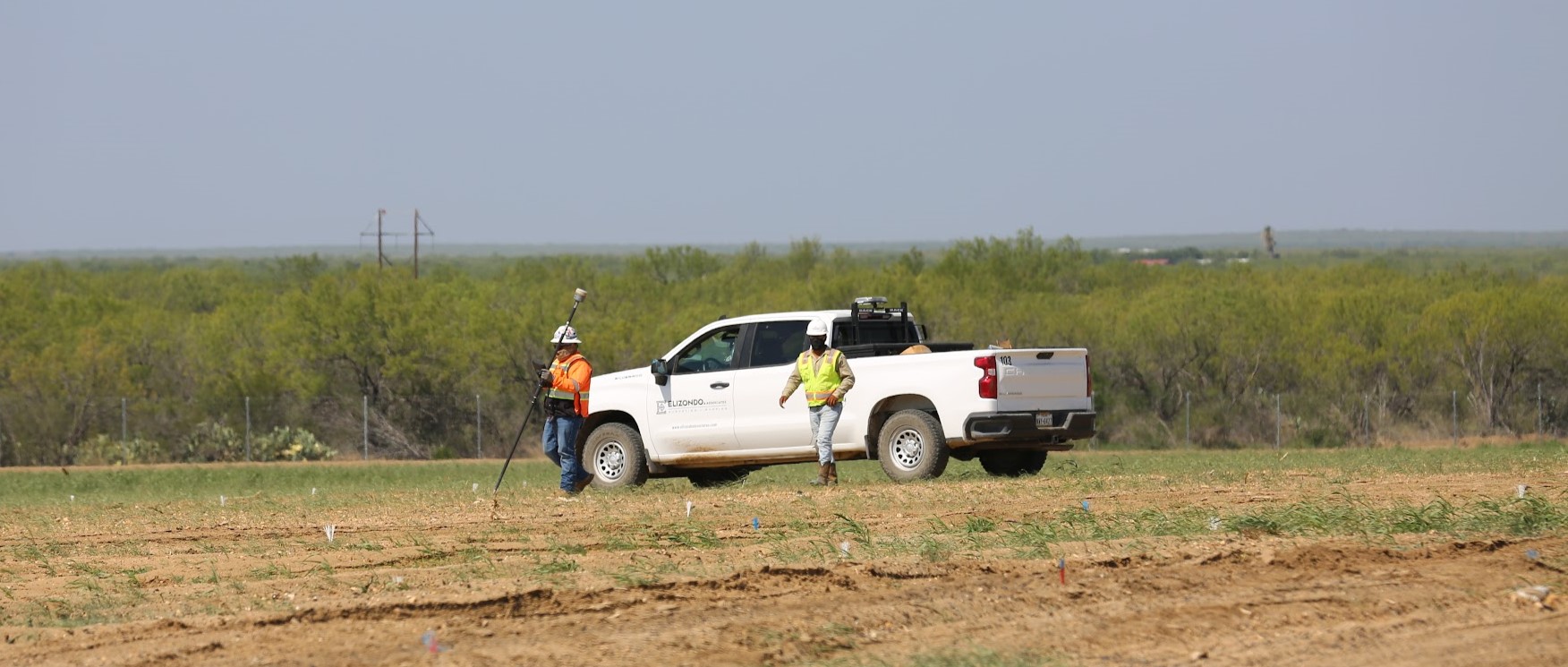 Two workers in safety gear using surveying equipment near a white pickup truck in a field.