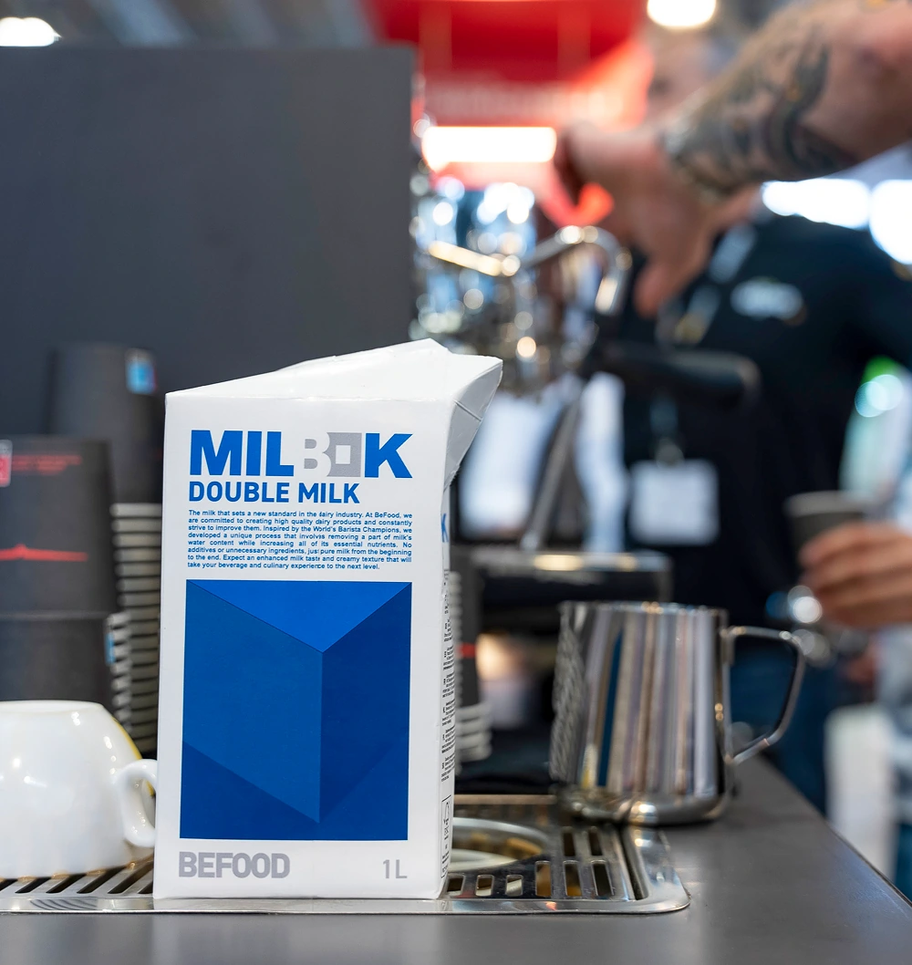 A 1-liter carton of MILBOX Double Milk, a specialty milk for cafés, sits on a coffee bar counter with a frothing jug, cups, and a person operating an espresso machine in the background.