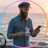 Bearded man in casual outfit holding coffee and smartphone near waterfront.
