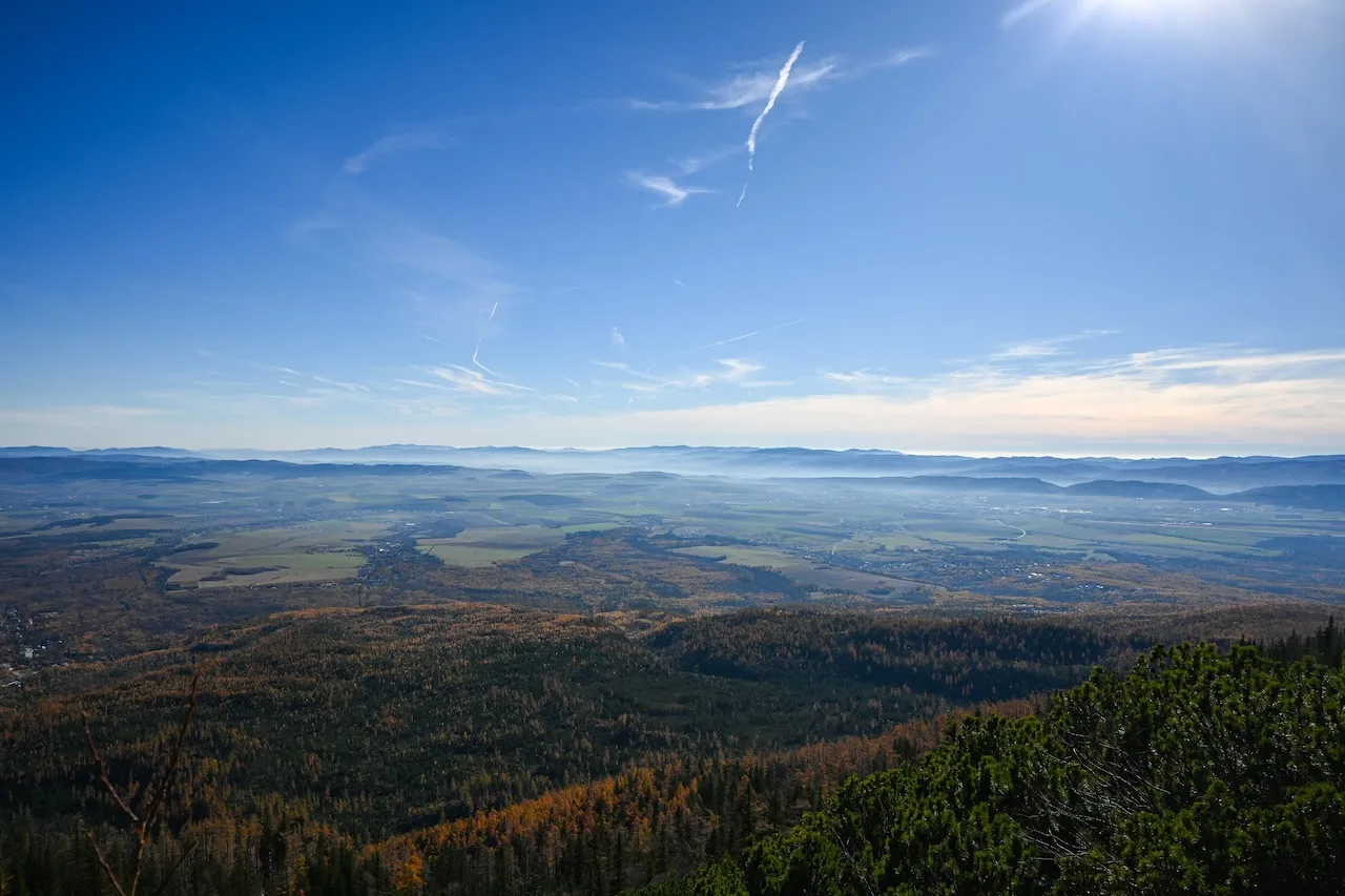 Panoramic view of a forested mountain valley with fields and distant hills under a clear blue sky with sun.