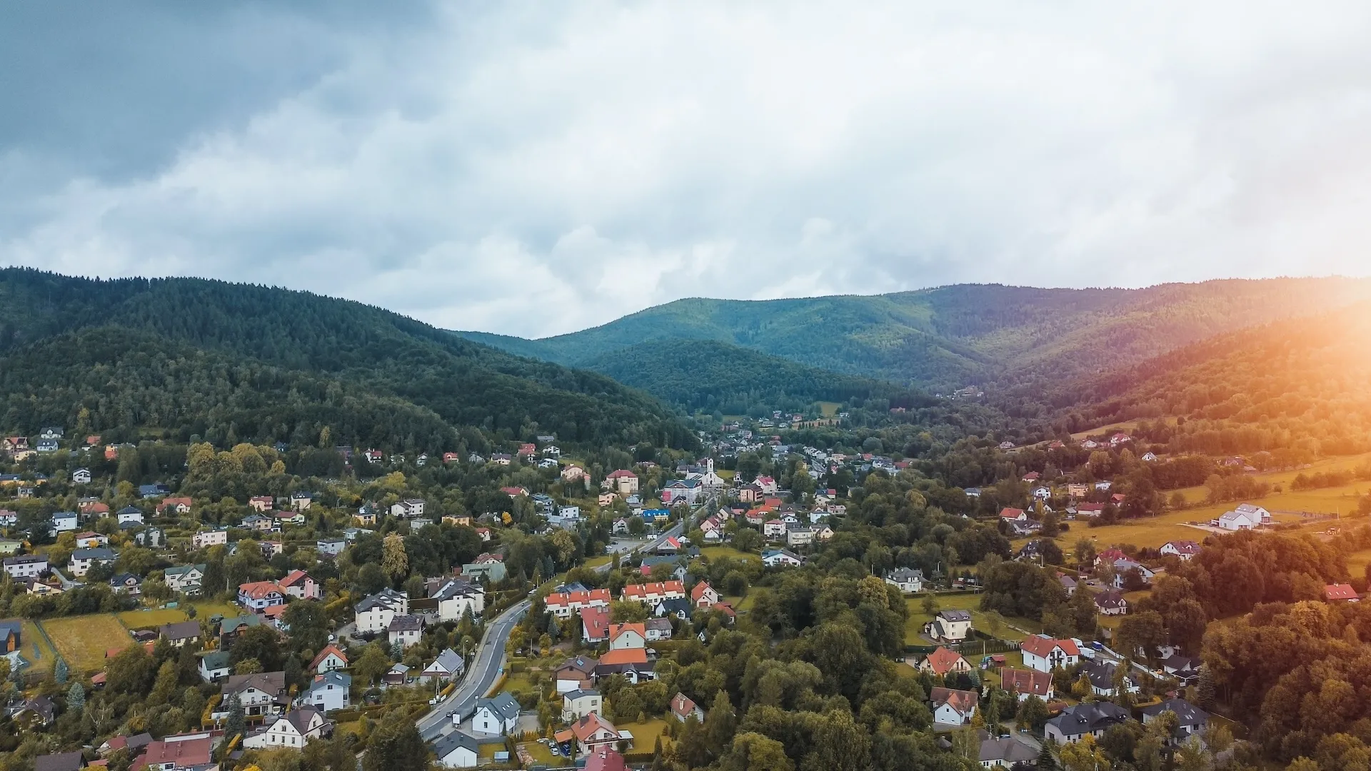 A scenic aerial view of a small town nestled in a lush green valley with forested hills under a cloudy sky and sunlight on the right side.