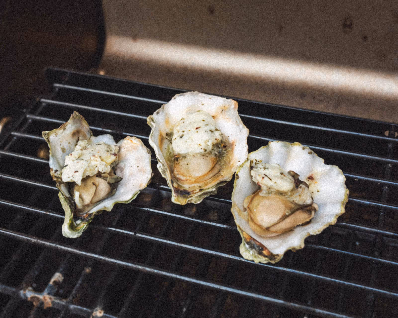 Three oysters sit open on the grates of a grill with a spoonful of garlic and herb compound butter melting on each of them