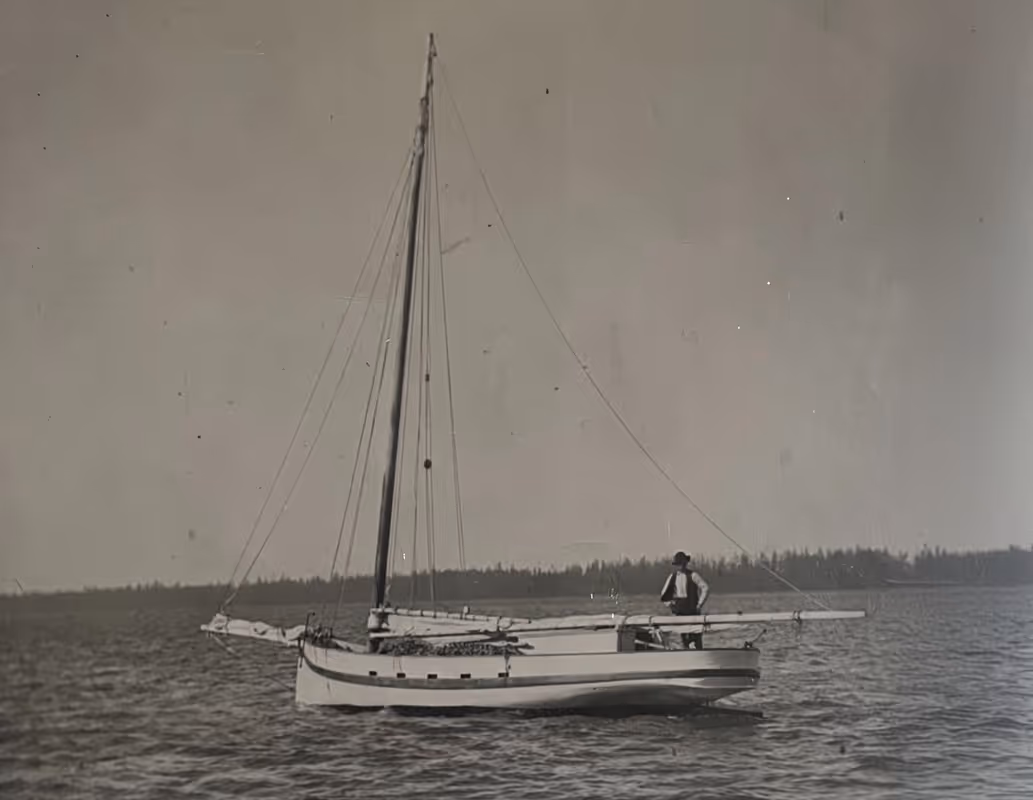 Black and white photograph of a large oyster boat with the sails down. An oysterman stands at the helm.