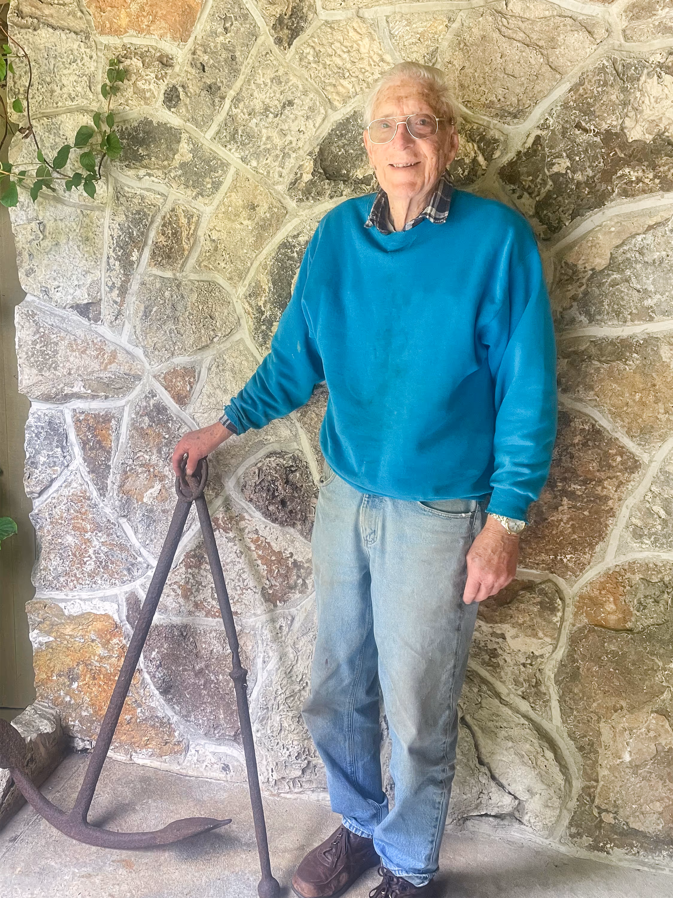 An older gentleman stands in front of an interior stone wall, wearing a teal crewneck and jeans, and resting his hand on an old, rusted anchor.