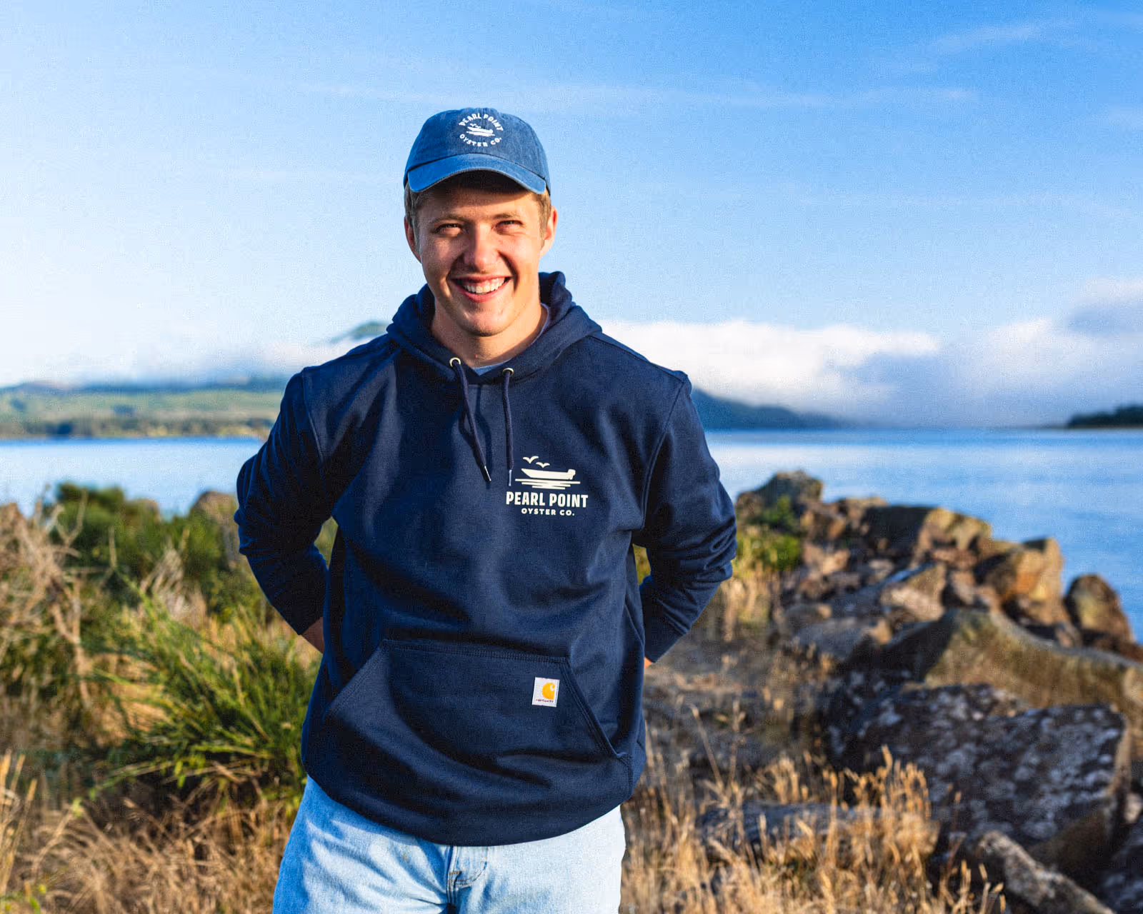 Max Weigardt, owner of Pearl Point Oyster Company, stands on a rocky jetty in Netarts Bay, smiling at the camera. He wears a denim hat and navy blue Carhartt sweatshirt, both with the Pearl Point Oyster Company logo on them.