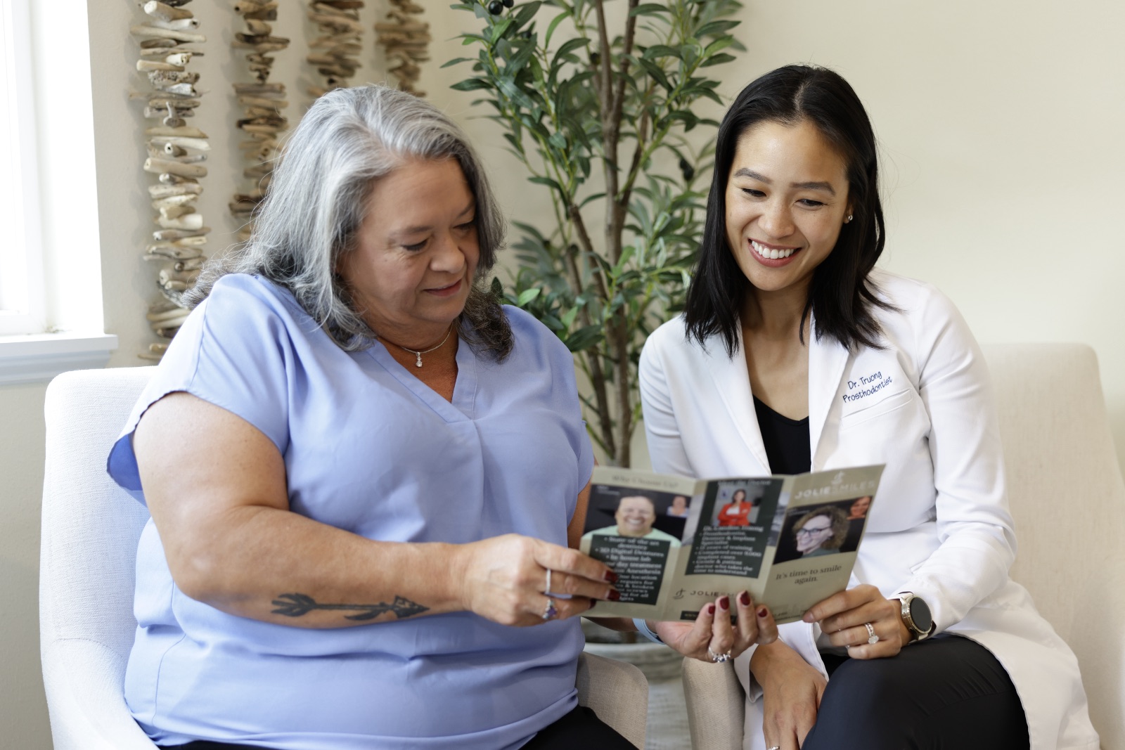 Dentist in white coat showing brochure to seated patient in blue shirt in a cozy office setting.