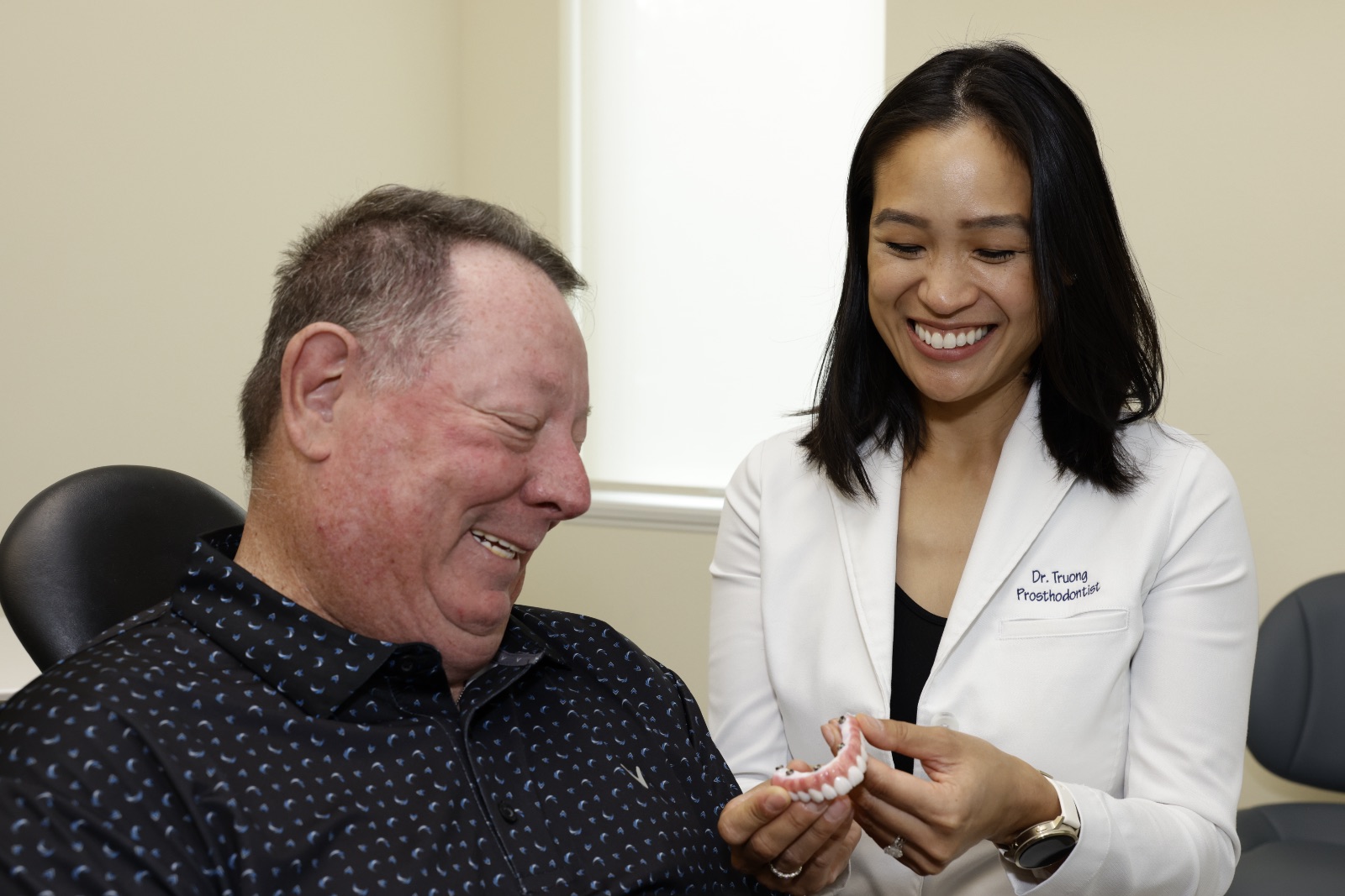 Smiling prosthodontist showing dental model to happy older male patient in clinic.