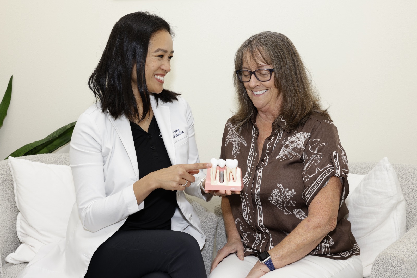 Dentist in white coat showing dental implant model to smiling woman with glasses.