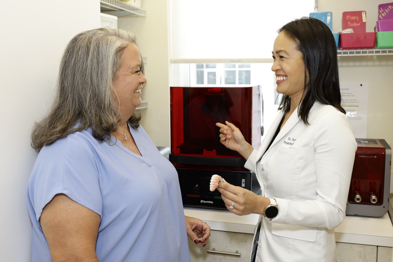Smiling dentist in white coat showing dental model to female patient in a clinic room.