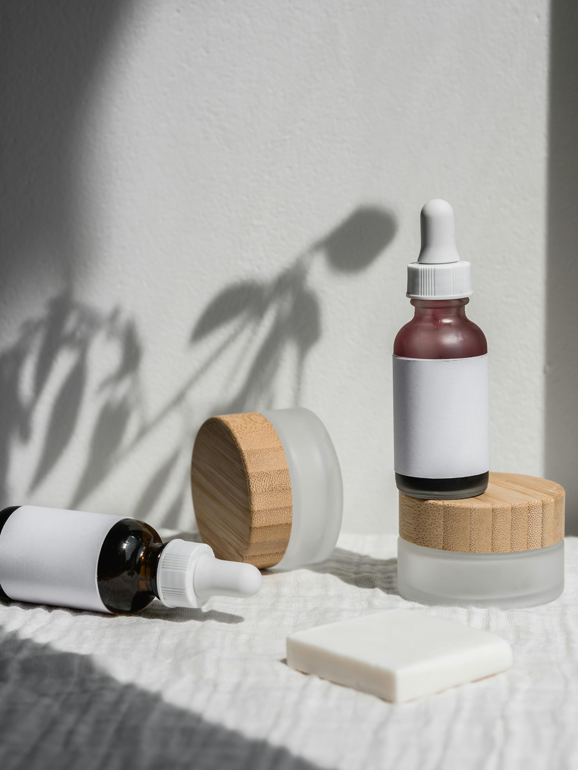 Two amber glass dropper bottles with blank white labels, two frosted glass jars with wooden lids, and a white soap bar on a textured white cloth with a plant shadow on the wall.