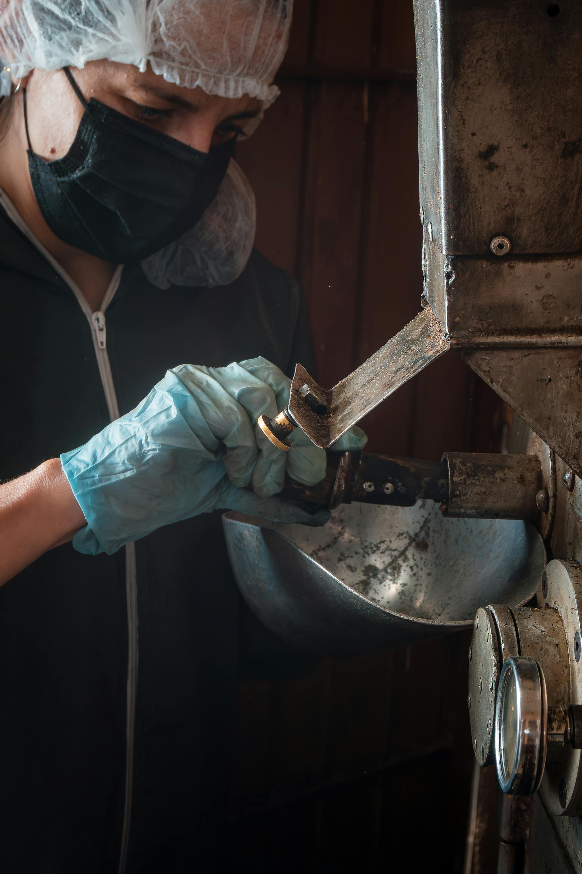 Person wearing a hair net, black face mask, and blue gloves adjusting a valve on industrial machinery.