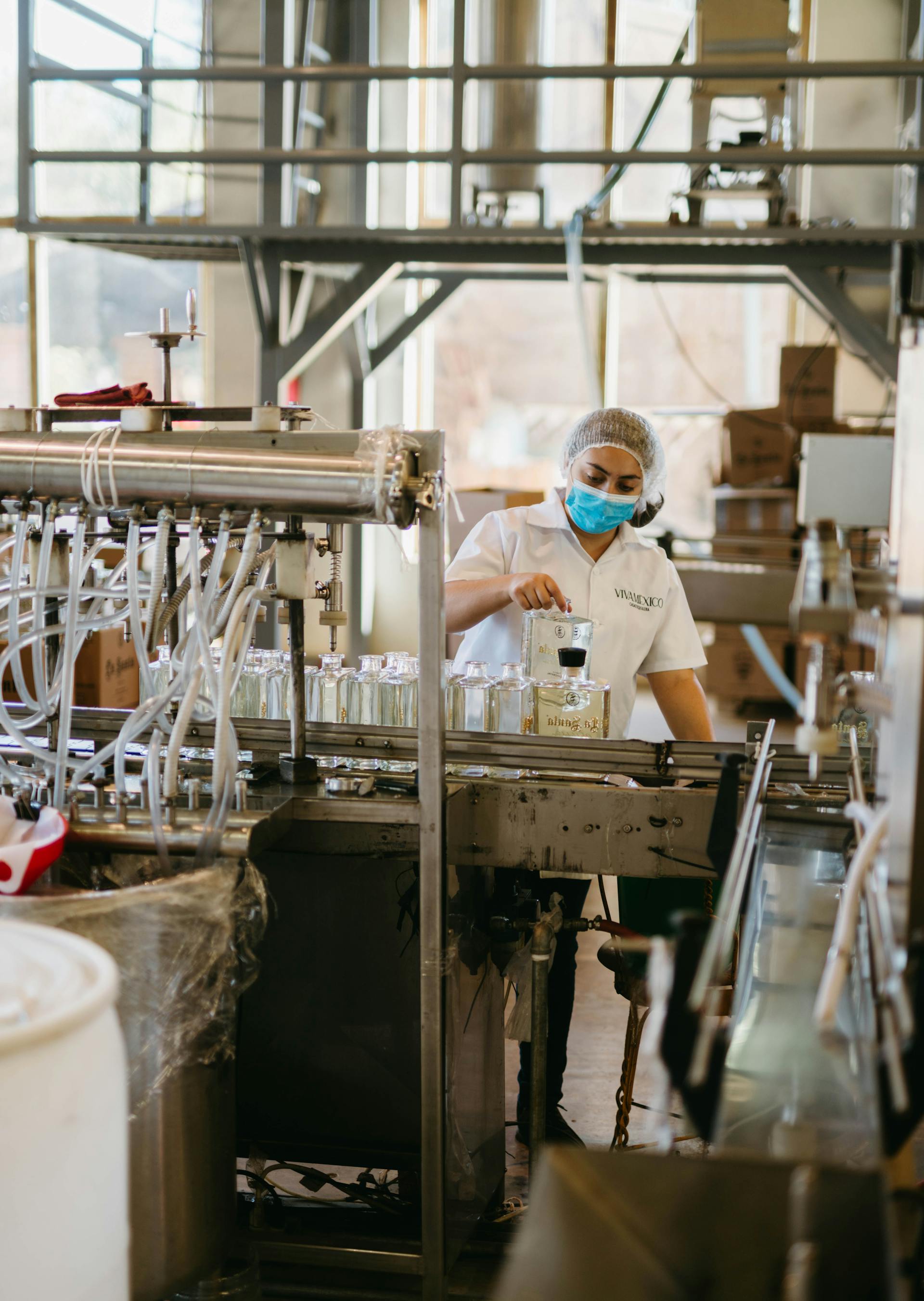 Factory worker wearing a hairnet and mask filling glass bottles on an automated production line.