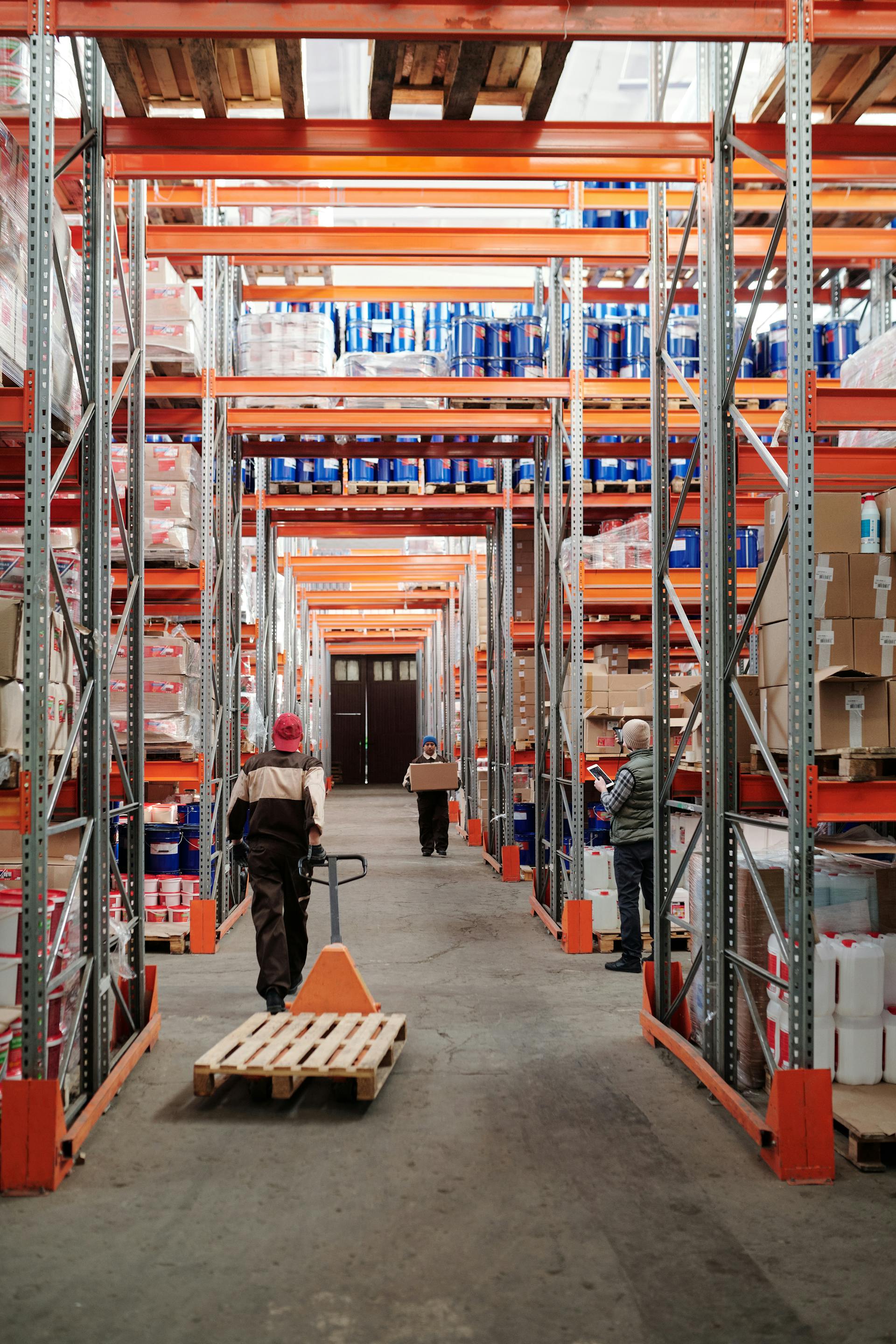 Three workers organizing and moving pallets in a large warehouse with metal shelves stocked with boxes and containers.