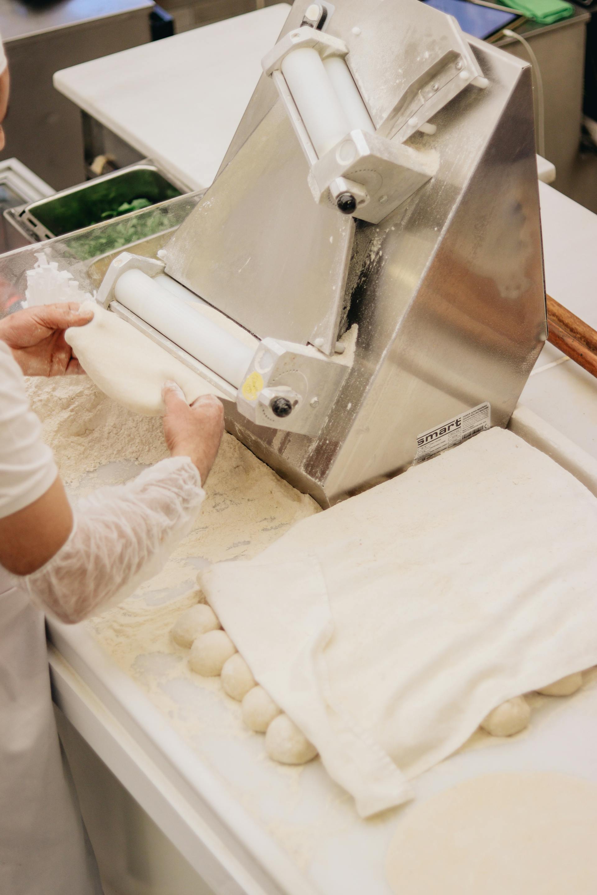 Person rolling dough using a stainless steel dough sheeter machine with dough balls covered by a cloth on a floured surface.