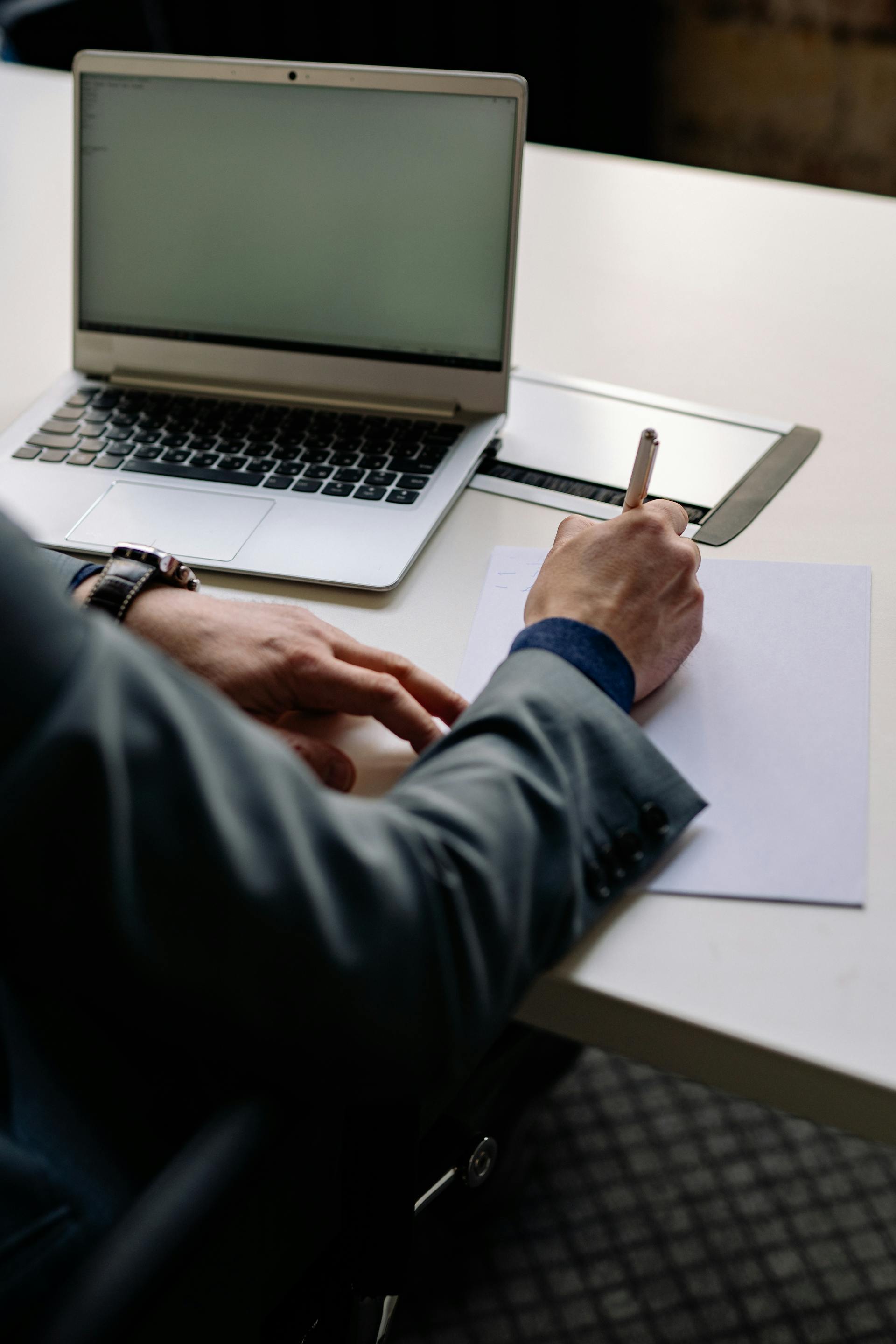 Person in business attire writing on a blank sheet of paper beside an open laptop on a white desk.