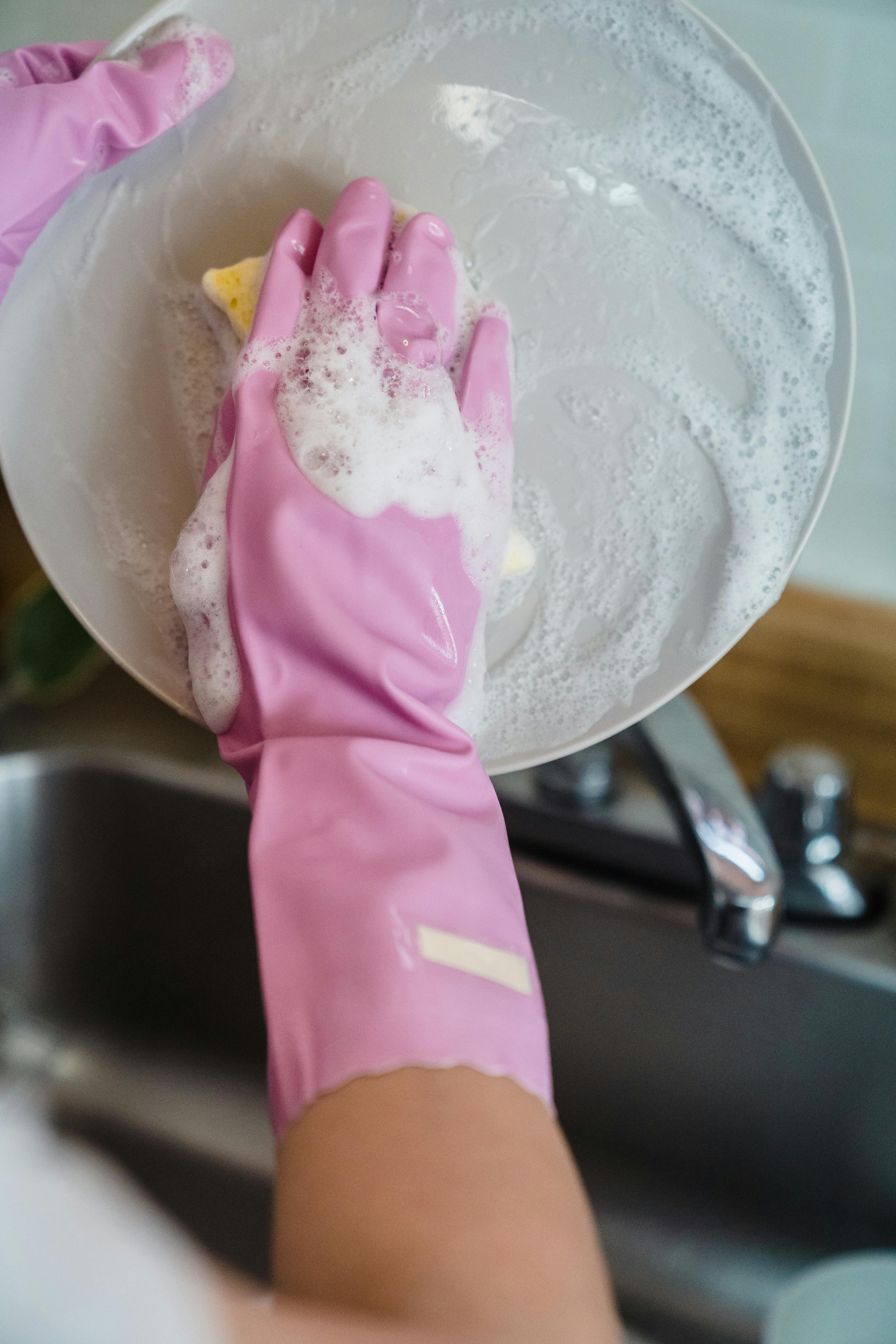 Person wearing pink gloves washing a white, soapy plate over a kitchen sink.