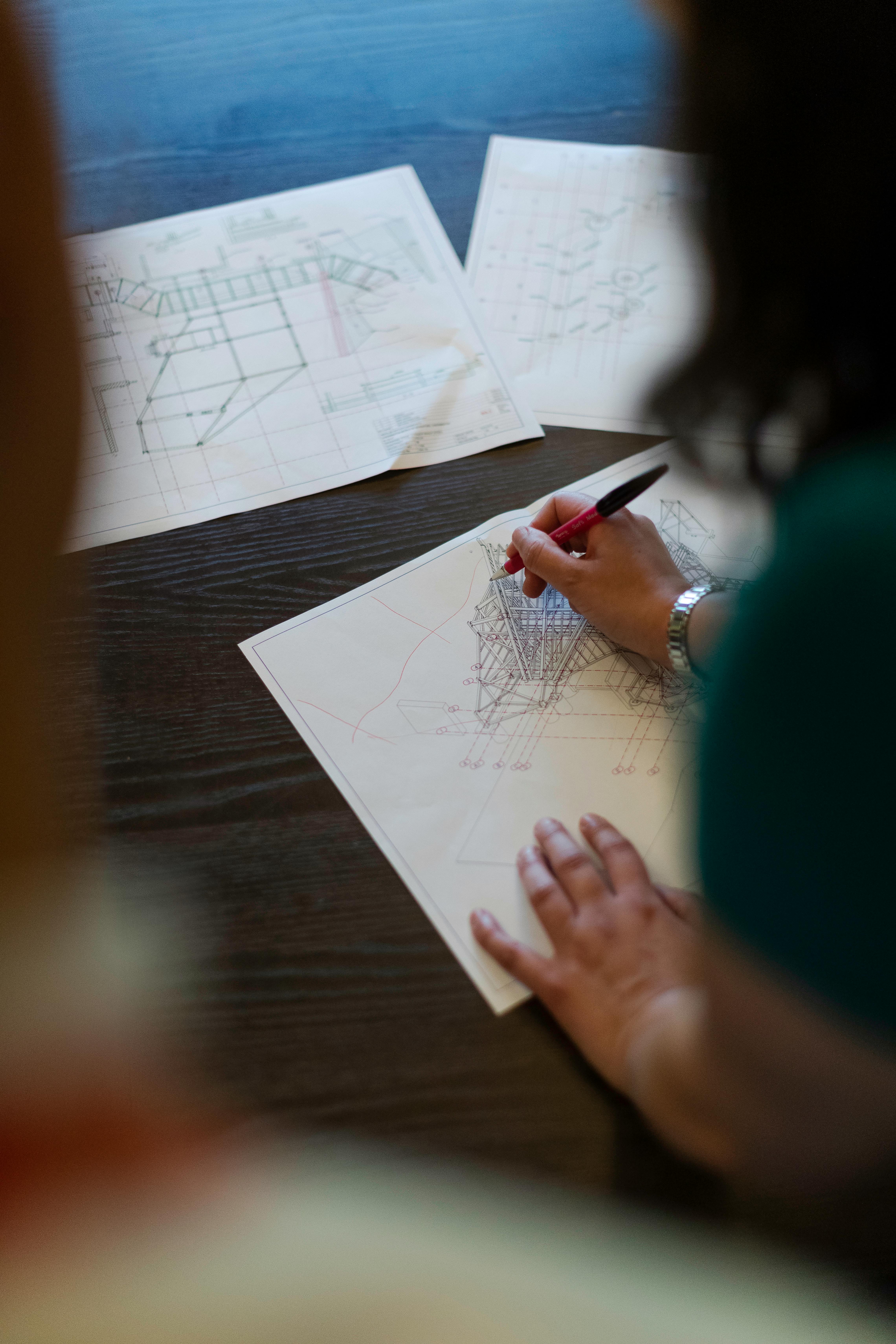 Person marking a technical drawing or architectural blueprint with a red pen on a dark wooden table.