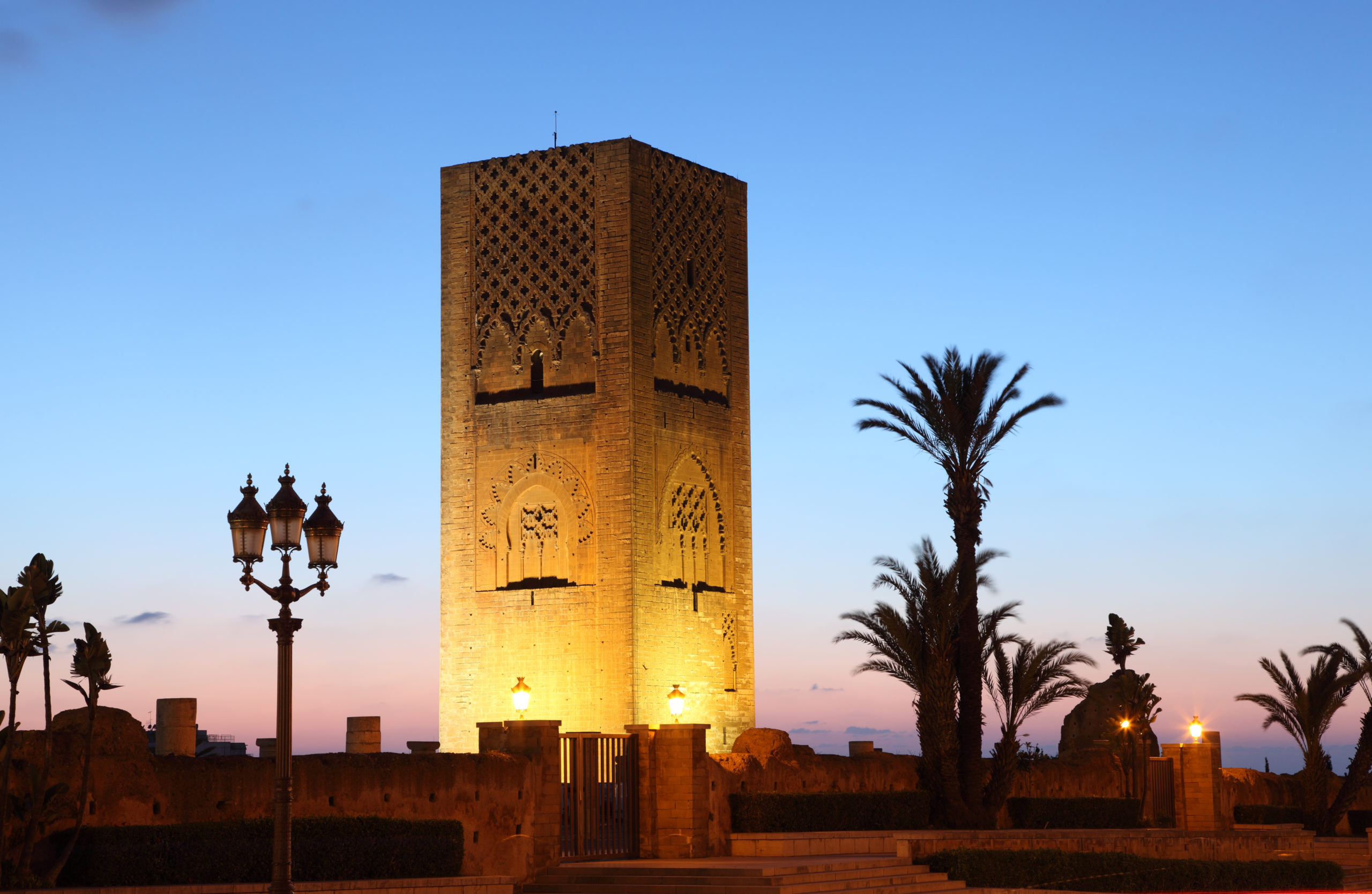 Historic Hassan Tower in Rabat a stone tower illuminated at dusk with intricate carvings, surrounded by palm trees and ornate street lamps under a clear blue sky fading into sunset during a tailor made tour in morocco