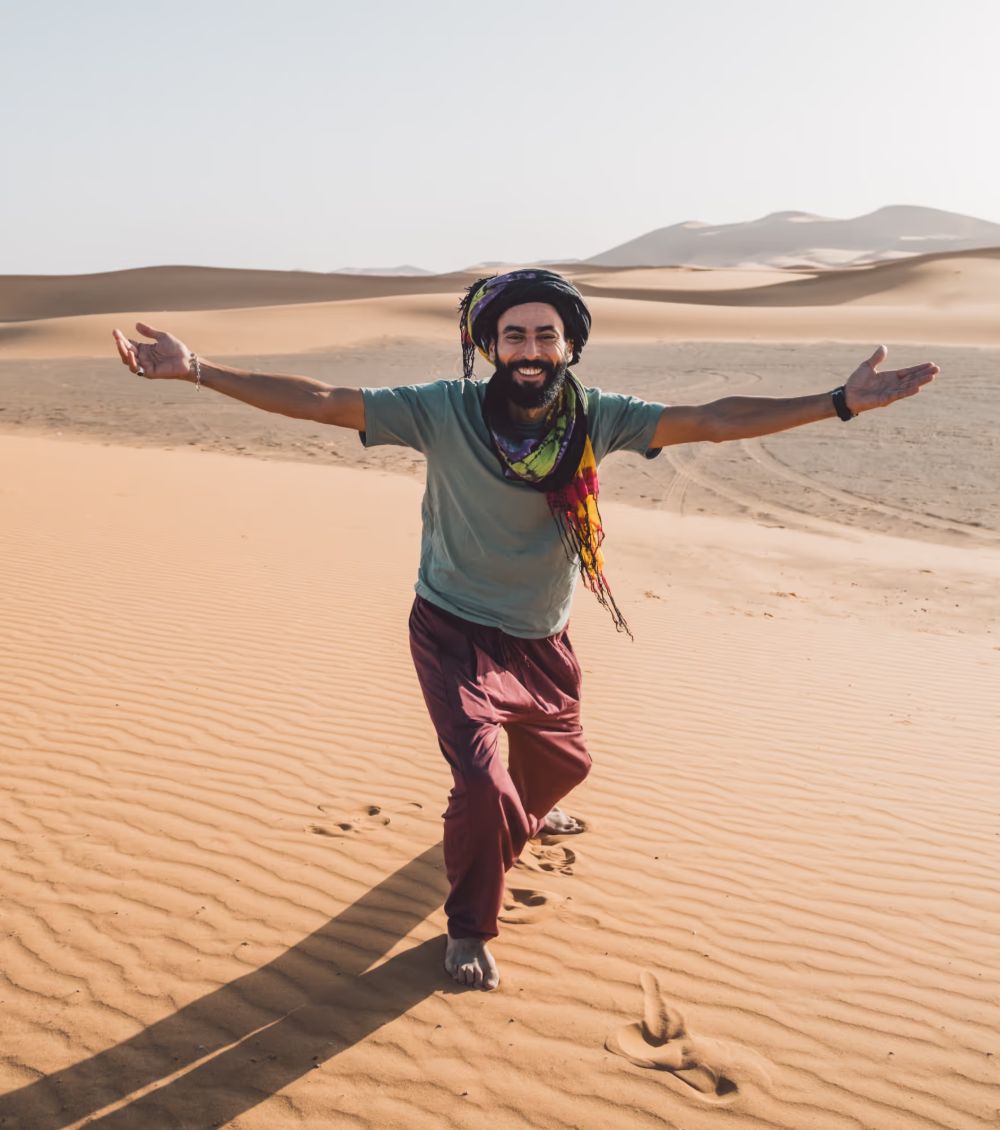Noureddine Ingrioui, founder of Moroccan Guides Travel, standing barefoot with his arms open on the dunes of the Sahara
