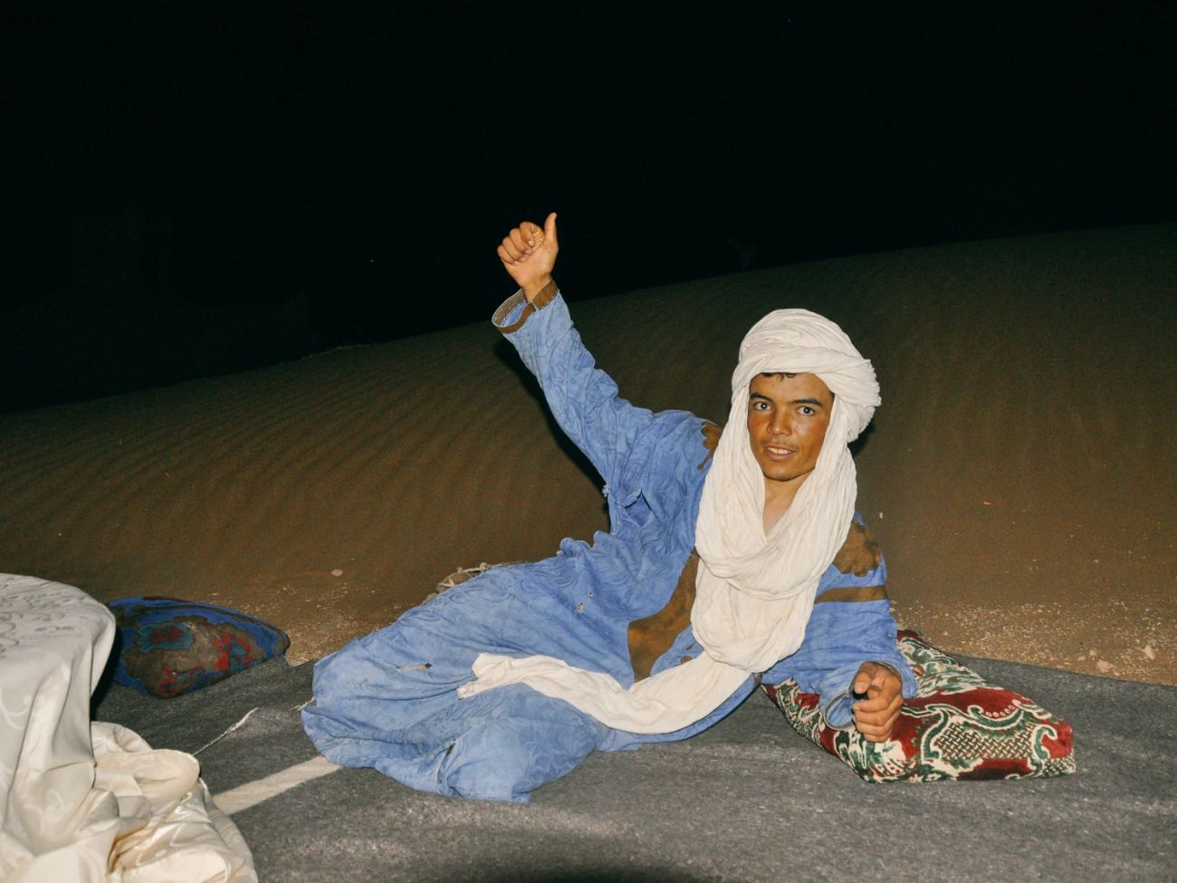 A young Moroccan guide sitting on a blanket in the desert at night, smiling at the camera
