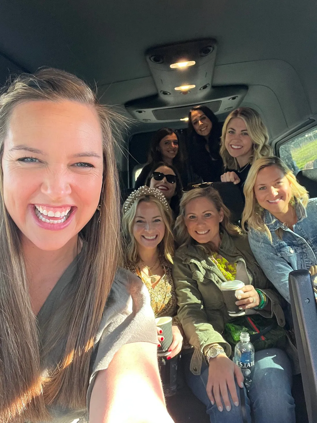 A group of women taking a selfie inside a van, smiling and holding drinks, during a trip through Napa Valley.