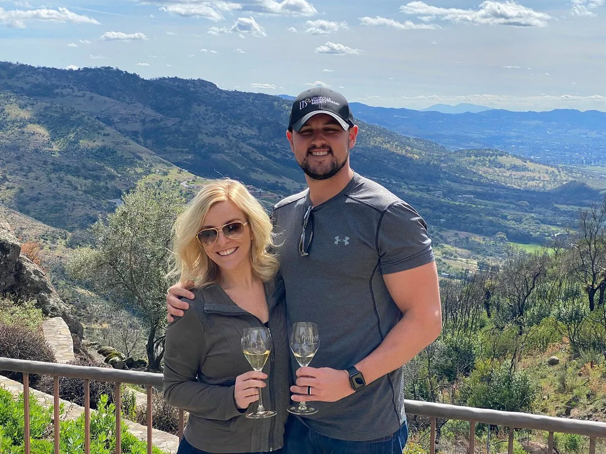 A couple standing together holding wine glasses, enjoying a scenic view of Napa Valley hills and vineyards from a balcony.