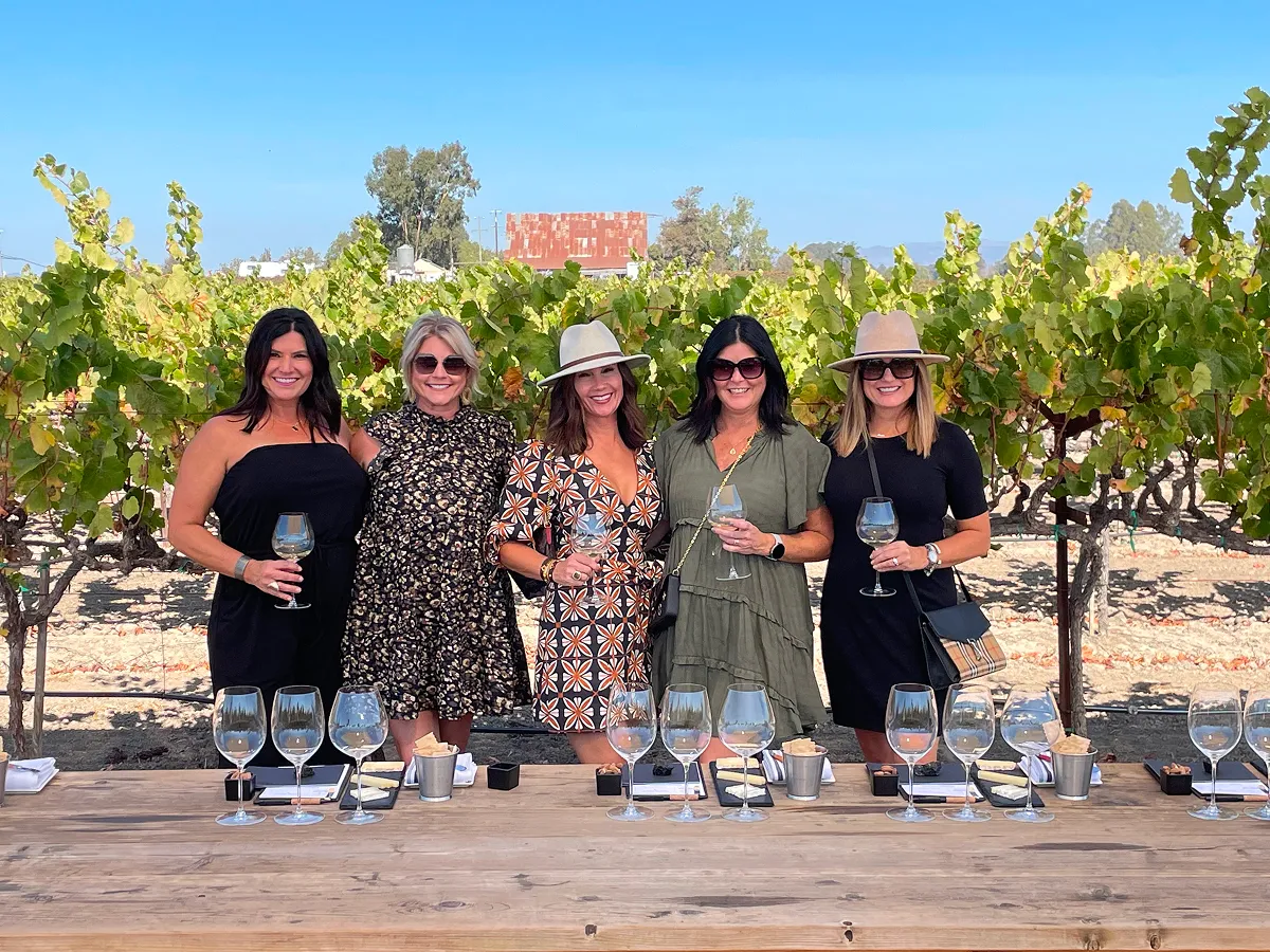 A group of five women standing behind a table with wine glasses, enjoying a wine tasting event in a Napa Valley vineyard with grapevines and a clear blue sky in the background.