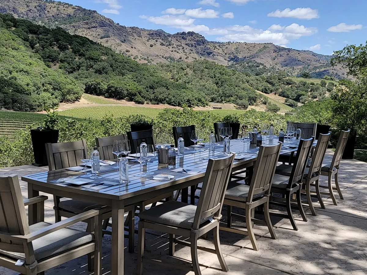 A long outdoor dining table set with chairs, glasses, and water bottles, overlooking Napa Valley vineyards and rolling hills under a clear blue sky.